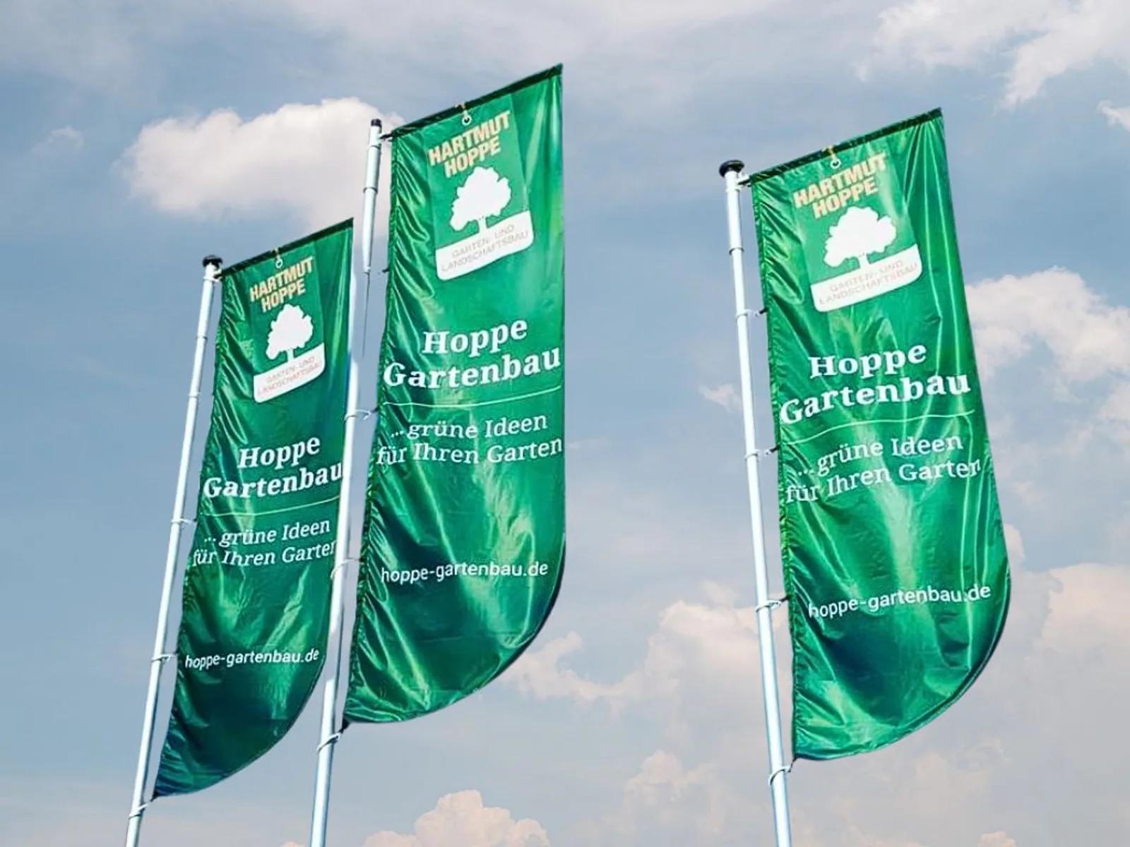 Drei grüne Werbefahnen von Hoppe Gartenbau mit dem Slogan „grüne Ideen für Ihren Garten“ vor blauem Himmel